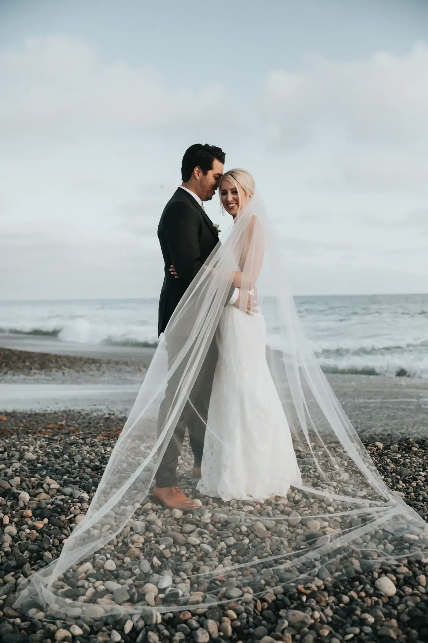 Couple embracing on a rocky Pacific coast beach, bride's cathedral veil sweeping in the ocean wind